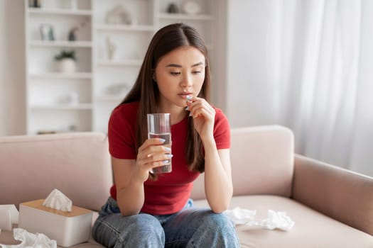 Concerned young asian woman taking pill with glass of water