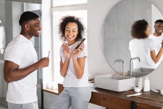 Cheerful Black Woman Shows Cellphone To Husband Cleaning Teeth Indoors