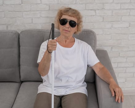 An elderly blind woman wearing sunglasses and with a tactile cane sits on the sofa.