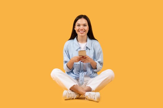 Happy european woman sitting with phone on yellow background
