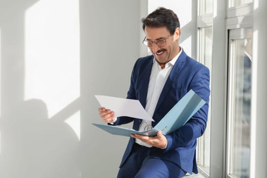 Cheerful businessman in a blue suit reads through documents in a bright office