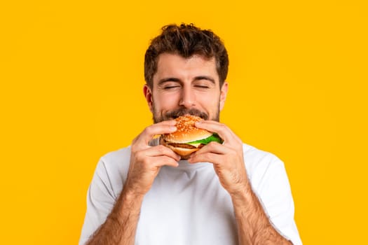 man enjoying overeating guilty pleasure eating burger over yellow background