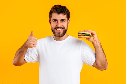 cheerful guy holding cheeseburger gesturing thumbs up on yellow background