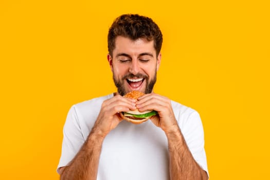 millennial guy enjoying tasty burger over yellow backdrop