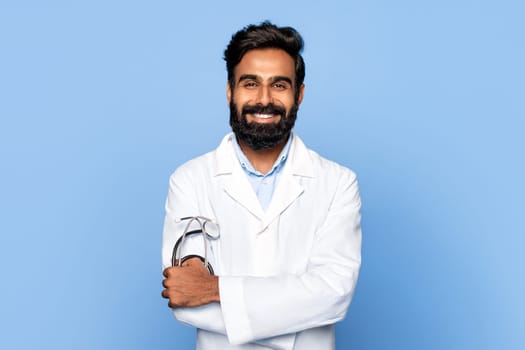 Smiling indian middle aged male doctor with crossed arms on blue backdrop