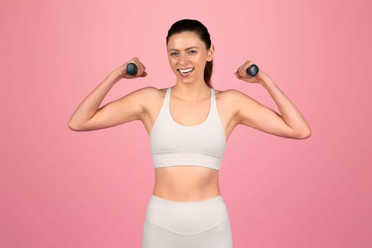 A cheerful woman flexes her muscles with a confident smile, holding small dumbbells
