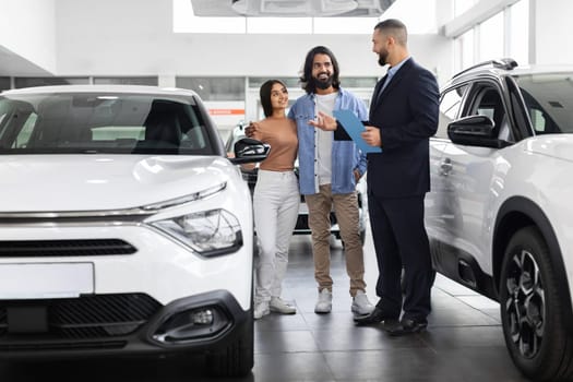 Attractive car salesman holding clipboard engages with prospective indian couple