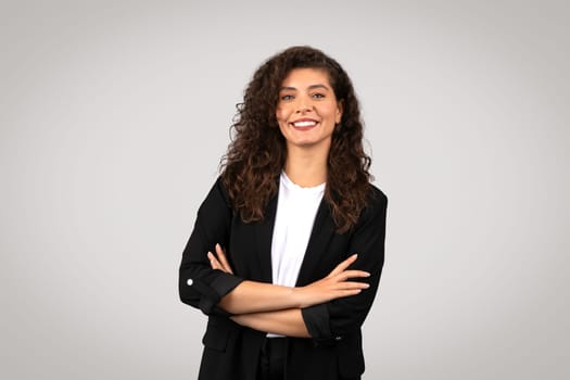 Cheerful young businesswoman in black blazer standing with folded arms and smiling against light grey background
