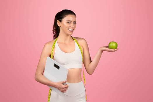 Cheerful fitness woman in a white sports outfit holds a green apple in one hand and a weighing scale