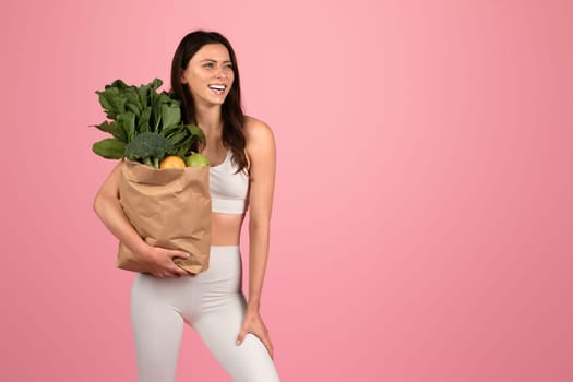 A joyful european woman in fitness attire laughs while holding a paper bag full of fresh vegetables and fruits, symbolizing healthy eating and lifestyle against a pink backdrop