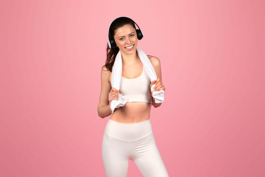 A smiling, athletic woman wearing a white sports bra and leggings with headphones, holding a towel over her shoulders