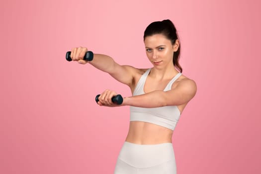 Focused and determined, a woman in a white sports bra and leggings holds out dumbbells with a steady gaze