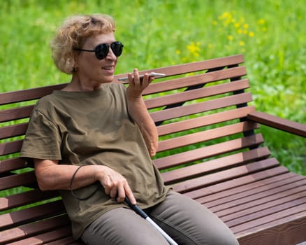 An elderly blind woman sits on a bench in the park and talks on a smartphone.
