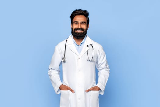 Portrait of confident male doctor with hands in pockets on blue backdrop