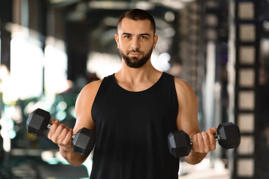 Gym Workout. Young Muscular Man Training With Dumbbells In Modern Sport Club, Portrait Of Motivated Confident Male Athlete Exercising With Light Weights And Looking At Camera, Copy Space