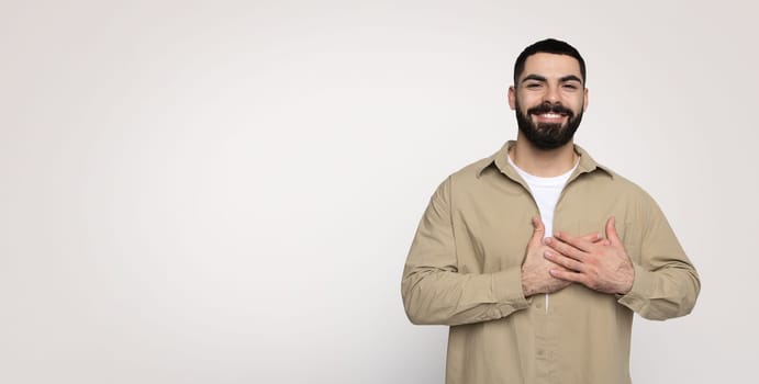 Glad confident handsome young middle eastern man with a beard in casual put hands to heart chest