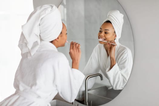 Cheerful black woman brushes teeth smiles to reflection in bathroom