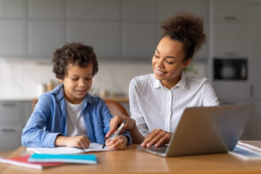 Black mother helps son with homework on laptop in well-lit kitchen