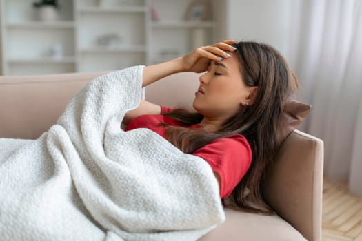 Exhausted asian woman touching her forehead, lying down on couch under blanket