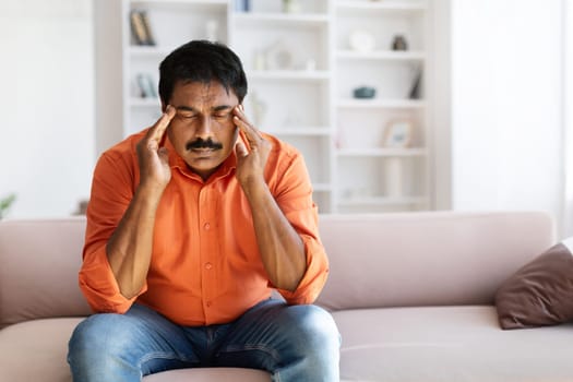 Stressed mature indian man sitting on couch, rubbing temples