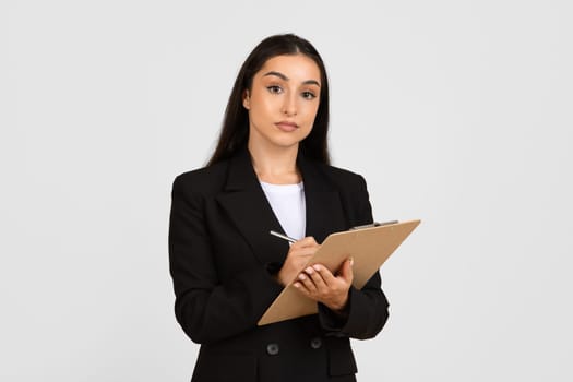 Serious businesswoman holding clipboard and writing, looking at camera