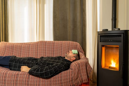Sick Hispanic man lying on the couch with a robe and a cloth on his forehead in the heat of a pellet stove