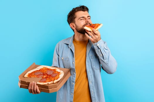 Hungry millennial man enjoys taste of pizza slice, blue backdrop