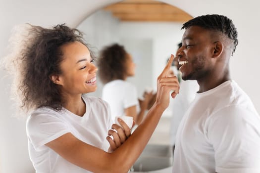 Playful Black Millennial Lady Applying Moisturizer On Husband's Nose Indoor