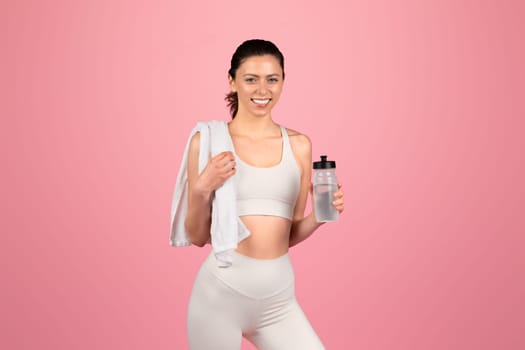 A happy, athletic woman in a white sports bra and leggings holds a water bottle and a towel over her shoulder