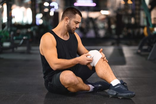 Man wrapping knee brace around his leg while sitting on gym floor