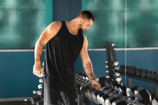 Athletic young man carefully selects dumbbells from the rack at gym