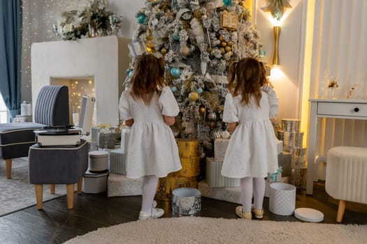 Two twin sisters in a white dress decorate the Christmas tree with their own hands, Christmas holidays at home.