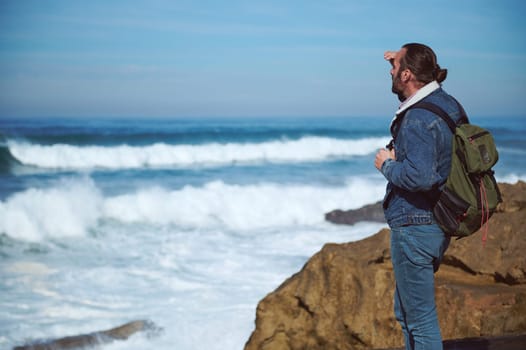 Absorbed bearded young male adventurer standing on a cliff and looking at horizon, enjoying the view of beautiful nature