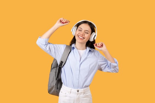 Joyful female student dancing with headphones on, yellow backdrop