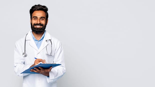 Smiling male doctor writing on clipboard, grey background, free space