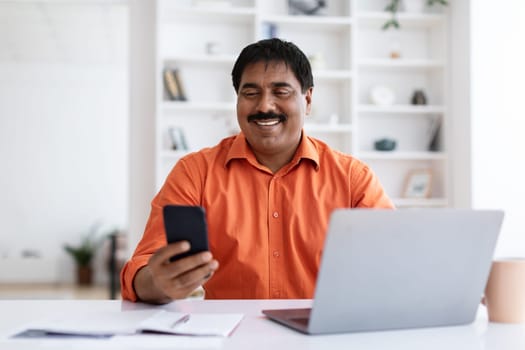 Positive middle aged hindu businessman using smartphone and laptop