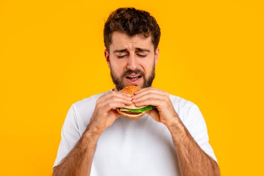 Unhappy bearded young man eats burger over yellow backdrop