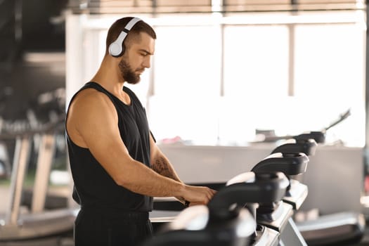 Jogging Workout. Young Muscular Man Wearing Wireless Headphones Using Treadmill At Gym