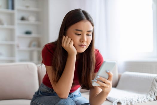 Pensive young asian woman looking at her smartphone with upset expression