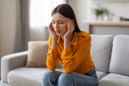 Distraught young woman with headache sitting on sofa at home