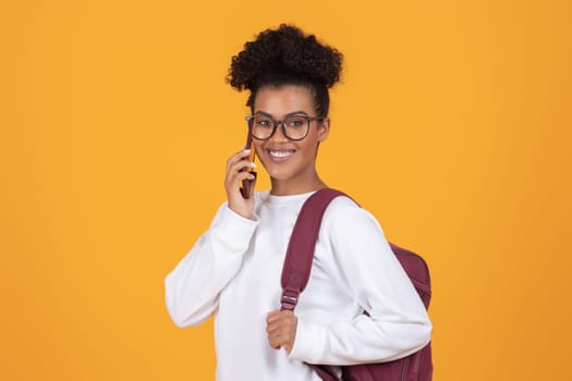 Smiling young black woman wearing glasses and backpack talking on cellphone