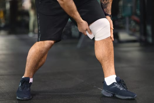 Sport Injury. Male athlete wrapping knee brace around his leg at gym