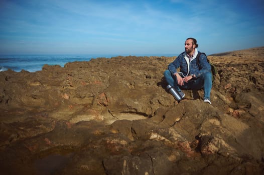 Young bearded man traveler, sitting alone on the rocky cliff, looking into the distance, enjoying the nature