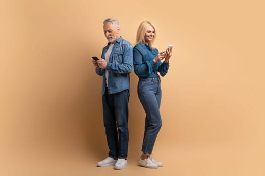 Smiling elderly couple use modern technologies, hold smartphones, reading text messages connected to wireless internet, wear casual denim outfits isolated over beige background. Retired pensioners