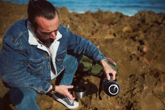 Bearded male tourist traveler pouring water from a touristic thermos into a cup, sitting on the rocky cliff in by ocean