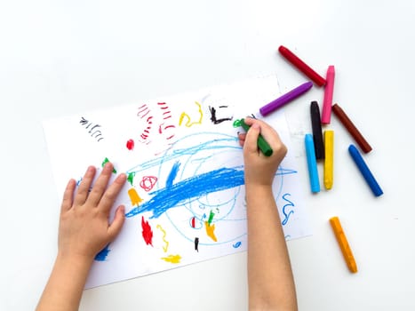Childs hands drawing with colorful wax crayons on white paper, top view. Creative art concept for educational and developmental activities.