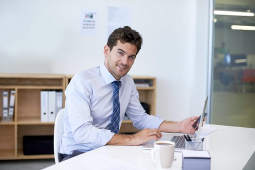 Portrait, desk and businessman with phone, laptop and coffee at startup with technology. Smile, online and business analyst in office with computer, smartphone and internet connection for networking