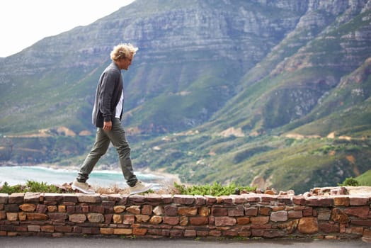 Travel, man and walking outdoor by mountain on holiday, vacation or trekking on trip in South Africa. Ocean, sea and person by stone wall for adventure, journey or tourist explore nature for hiking.