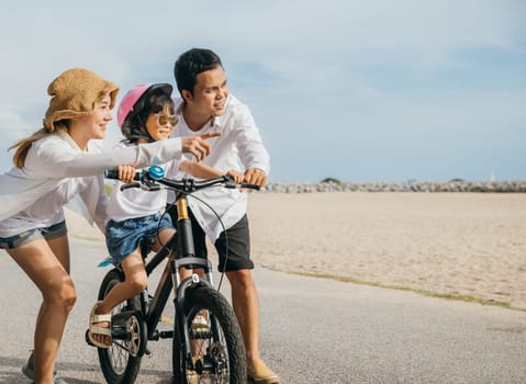 Family happiness unfolds on sandy beach as parents teach their children to ride bicycles during summer road trip. Smiles safety helmets and warmth of sun define this cheerful and carefree moment.