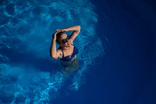 An elderly woman in sunglasses swims in the pool. Vacation in retirement.